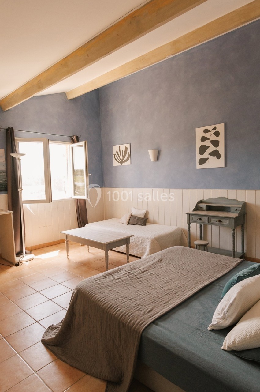 Chambre lumineuse avec deux lits, murs bleus, poutres apparentes, bureau et fenêtre ouverte sur l'extérieur.