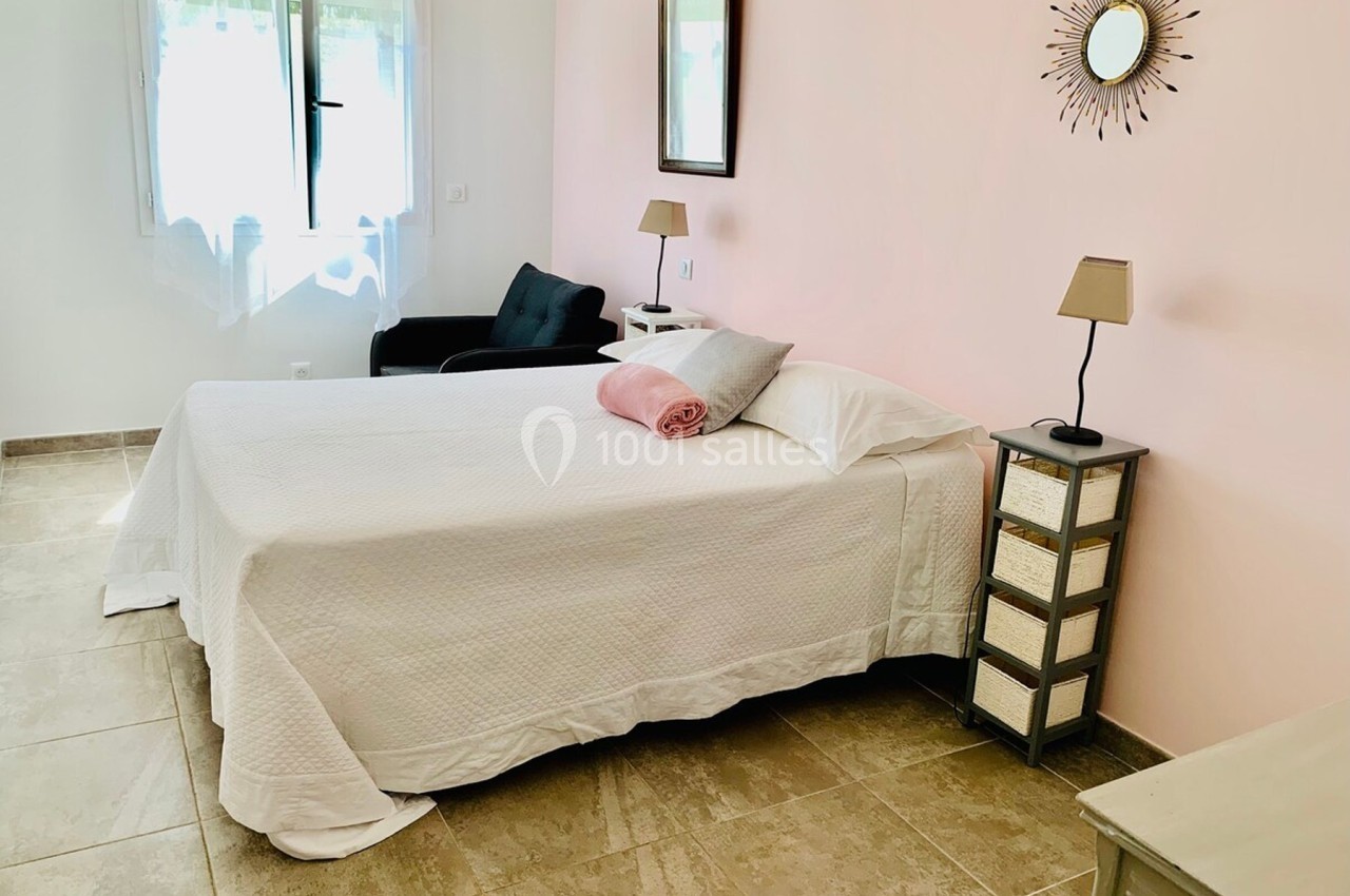 Chambre lumineuse avec un lit double, draps blancs, coussin rose, deux lampes de chevet et un mur rose clair.