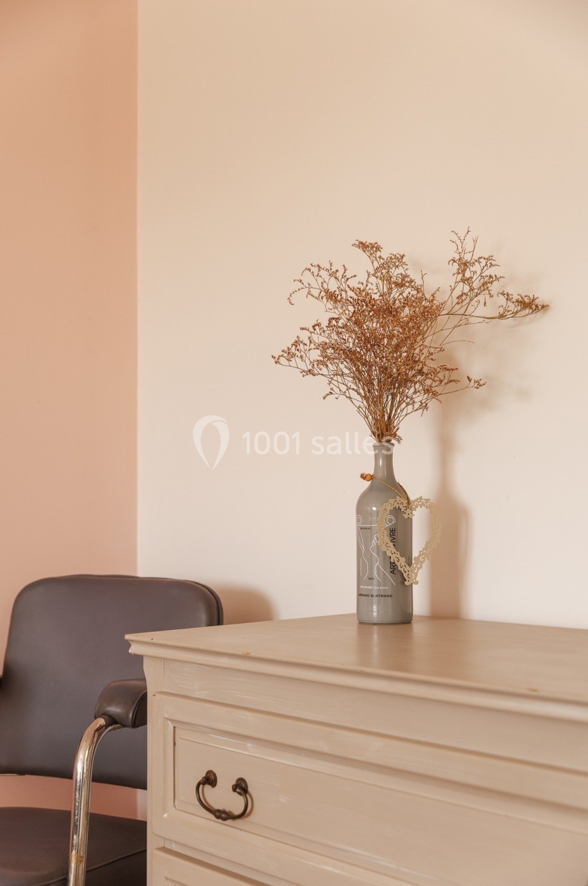 Un vase gris avec des branches séchées posé sur une commode beige, à côté d'une chaise noire.