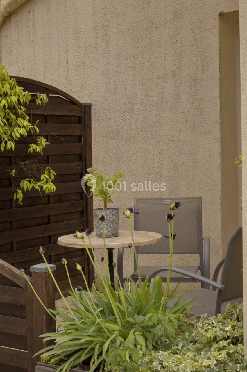 Petit coin terrasse avec table ronde, deux chaises, plantes en pot et iris en fleurs devant une clôture en bois.