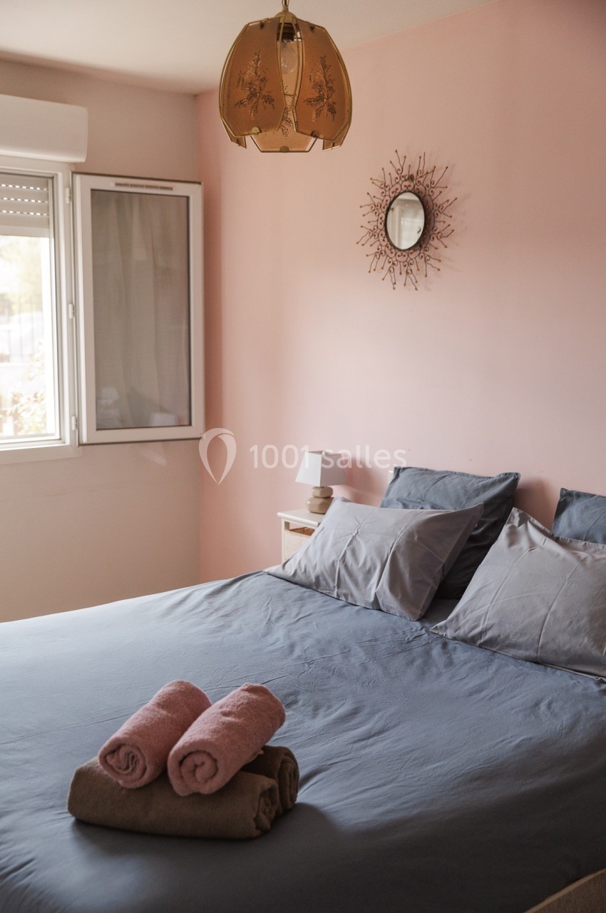 Chambre avec lit double, draps gris, serviettes pliées, mur rose, fenêtre ouverte et lampe suspendue.