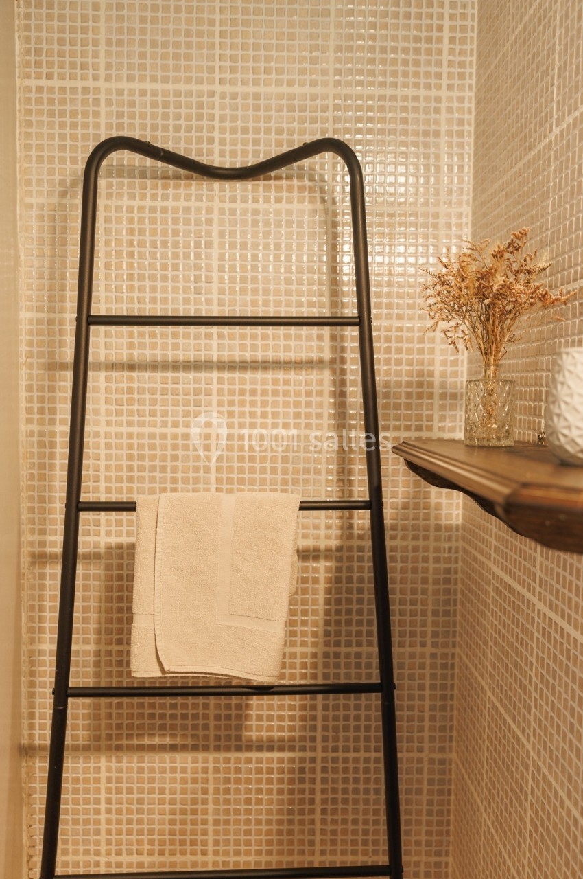 Échelle noire servant de porte-serviettes dans une salle de bain aux murs carrelés beige, avec une étagère en bois.
