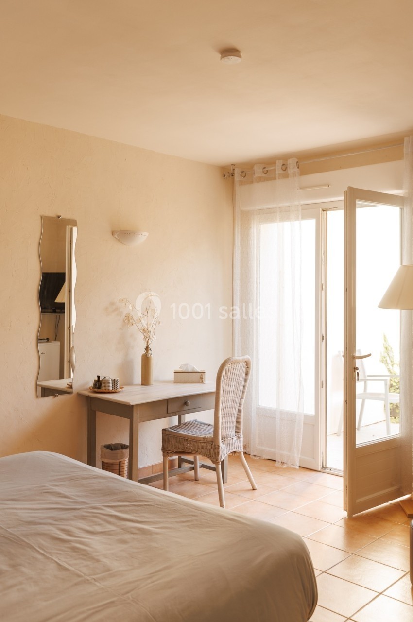 Chambre lumineuse avec bureau, chaise en rotin, lit, et porte-fenêtre ouverte sur un balcon.