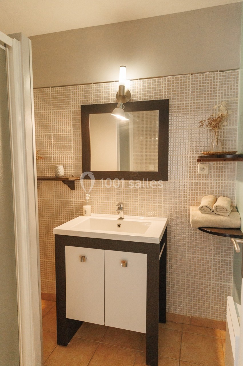 Lavabo moderne avec miroir encadré, éclairage mural, étagères décorées et carrelage beige au mur.