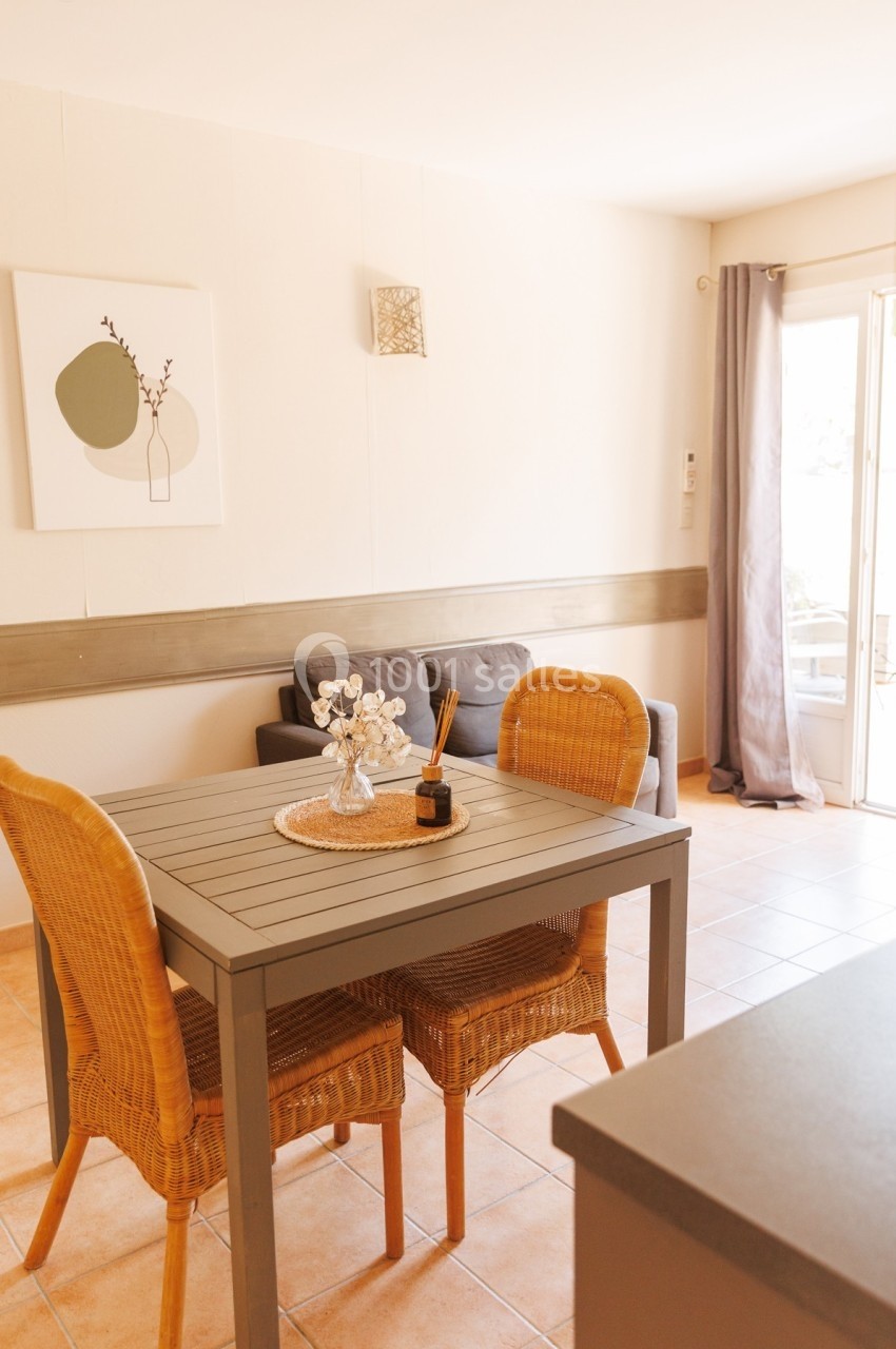 Salle à manger lumineuse avec table en bois, chaises en rotin, canapé gris et porte-fenêtre donnant sur l'extérieur.