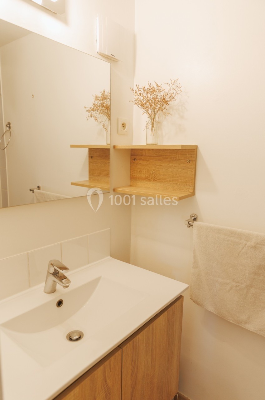 Lavabo blanc avec meuble en bois, miroir, étagères assorties et serviette suspendue dans une salle de bain lumineuse.