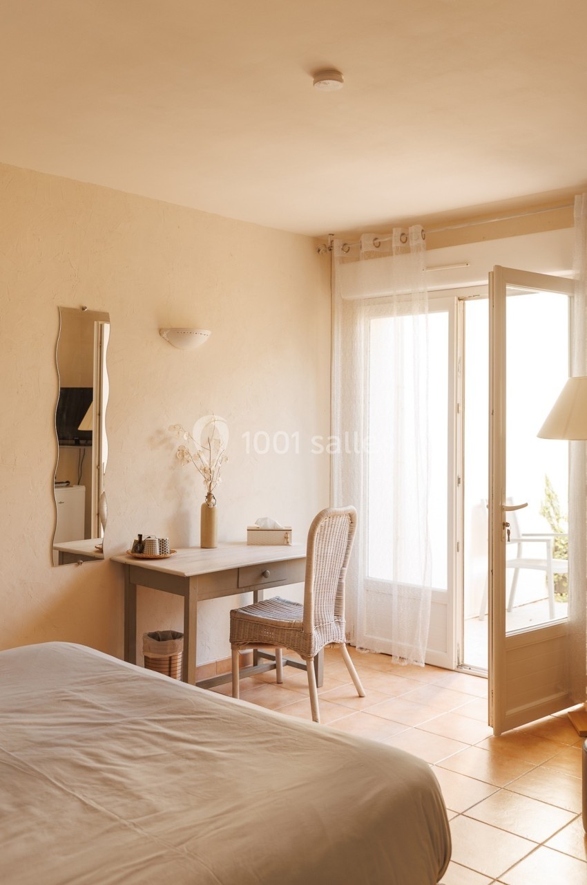 Chambre lumineuse avec bureau, chaise en rotin, lit, et porte-fenêtre ouverte sur un balcon ensoleillé.