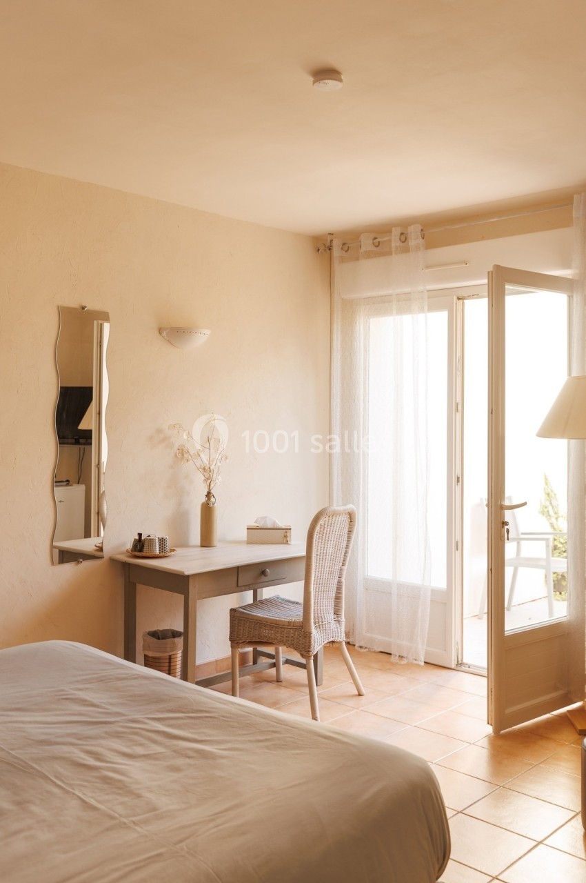 Chambre lumineuse avec bureau, chaise en rotin, lit, et porte-fenêtre donnant sur un balcon.
