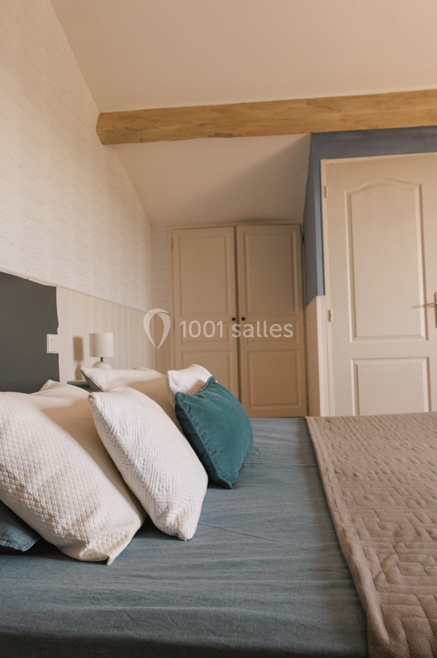 Lit avec draps bleus, coussins blancs et verts, dans une chambre lumineuse avec murs clairs et poutre en bois apparente.