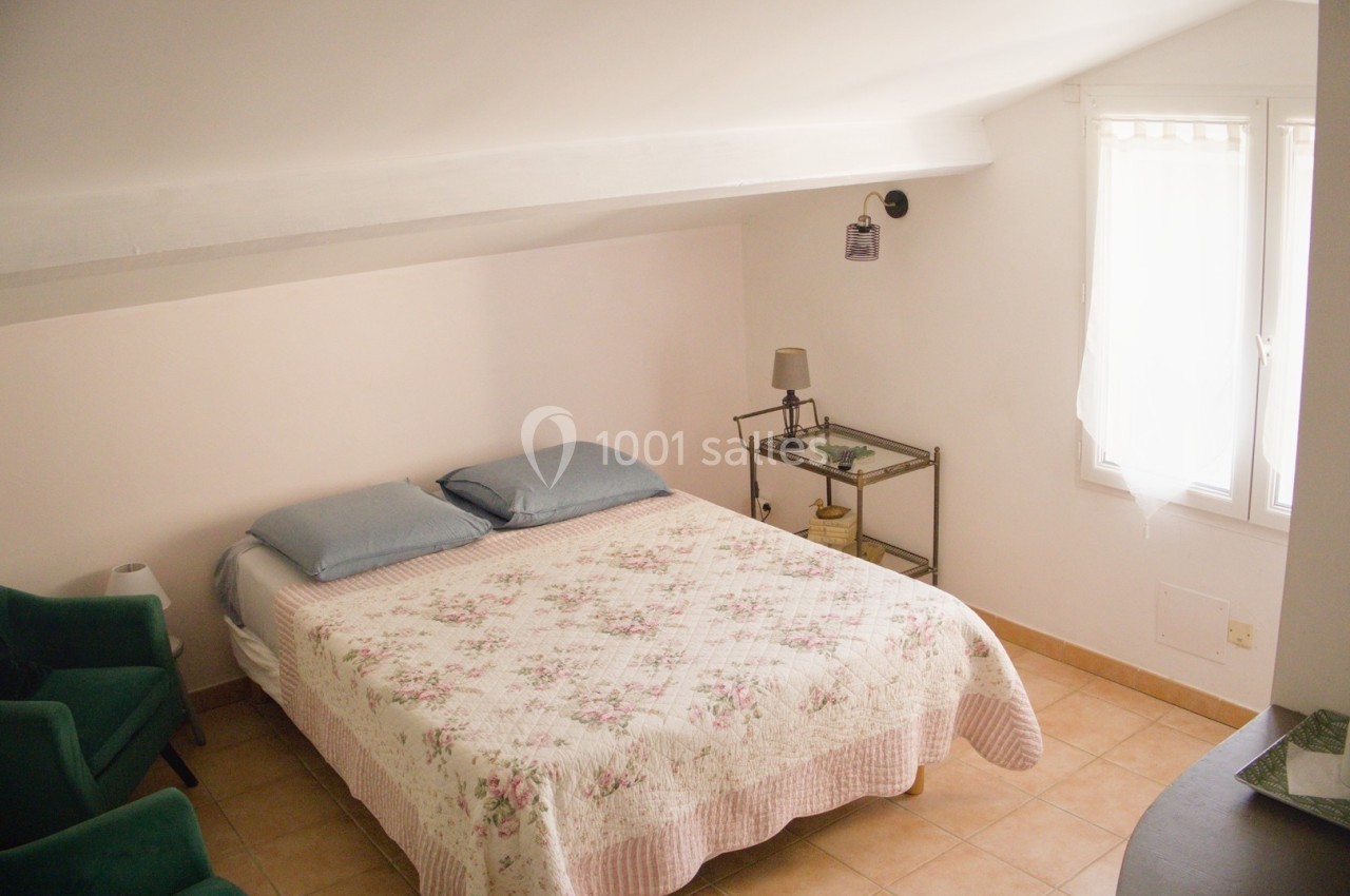 Chambre lumineuse sous combles avec lit double, fauteuil vert, table de chevet et fenêtre avec rideaux blancs.