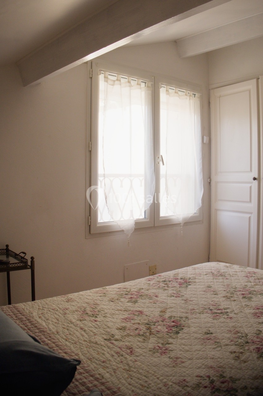 Chambre lumineuse avec un lit à couvre-lit fleuri, fenêtre à rideaux blancs et armoire intégrée blanche.