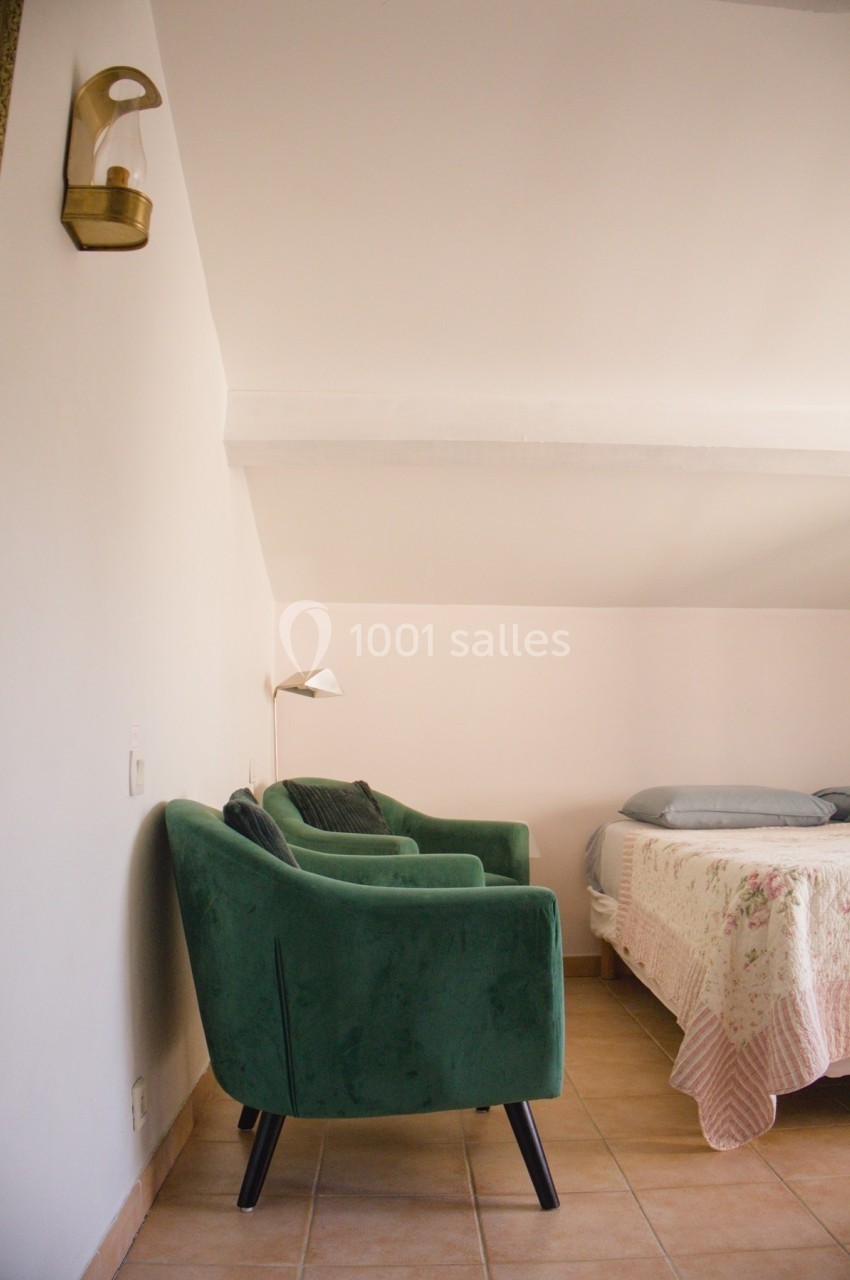 Fauteuil vert et lampe murale dans une chambre lumineuse avec lit double et couvre-lit fleuri.