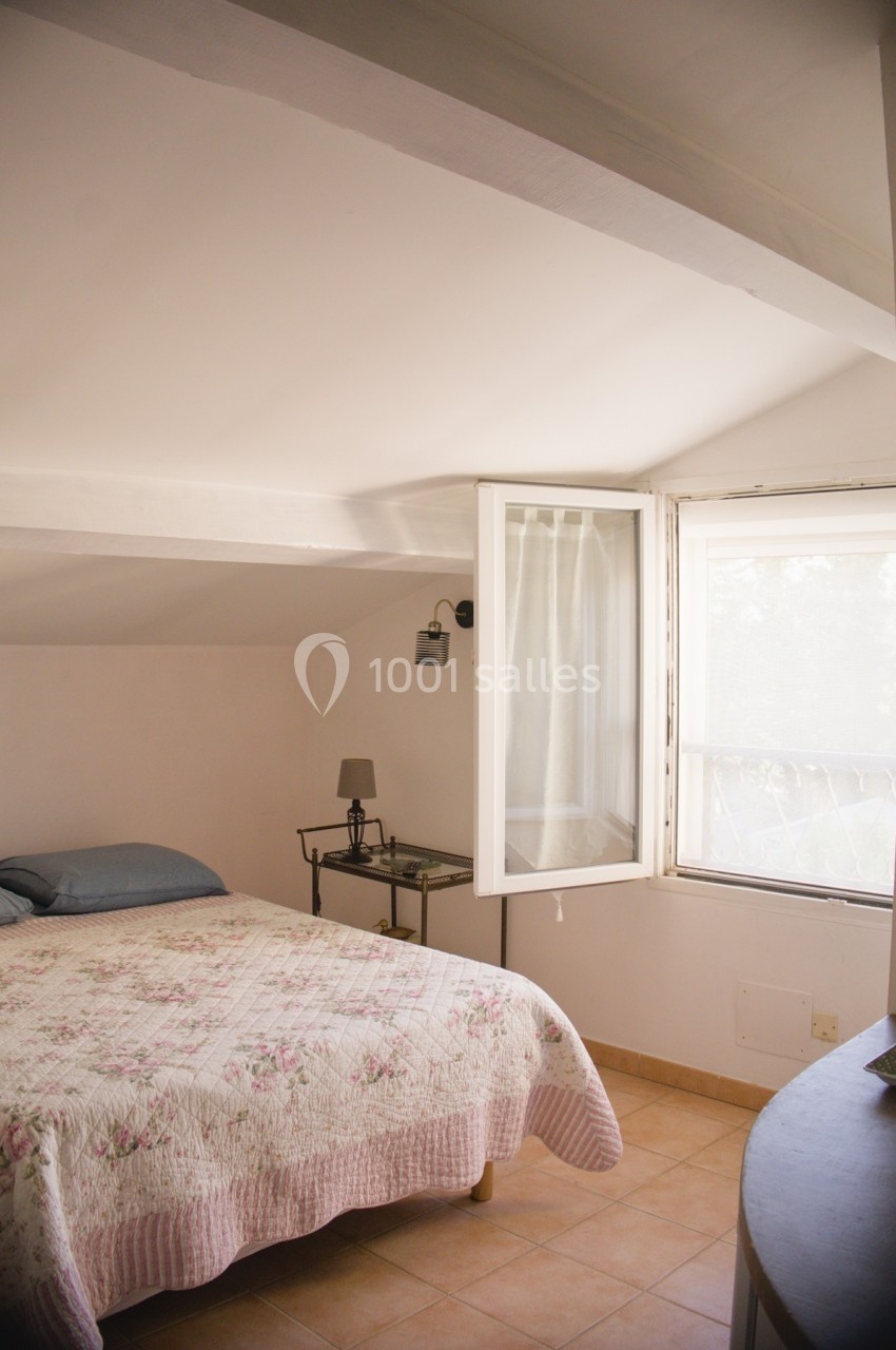 Chambre mansardée lumineuse avec un lit double, une table de chevet et une fenêtre ouverte laissant entrer la lumière…