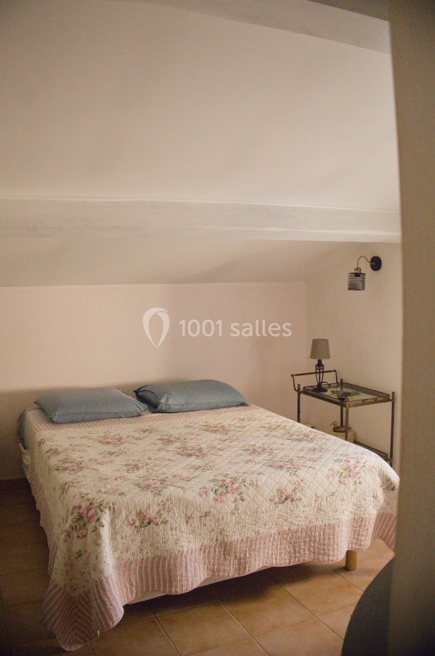 Lit double sous un plafond mansardé, couvert d'une couette à motifs floraux, avec table de chevet et lampe.