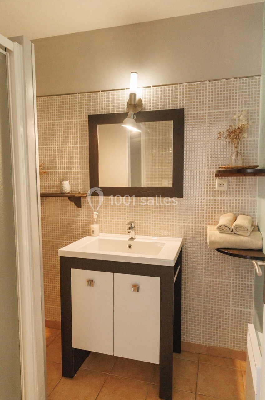 Lavabo blanc avec meuble en bois sombre, miroir encadré, éclairage mural et étagères décorées dans une salle de bain.