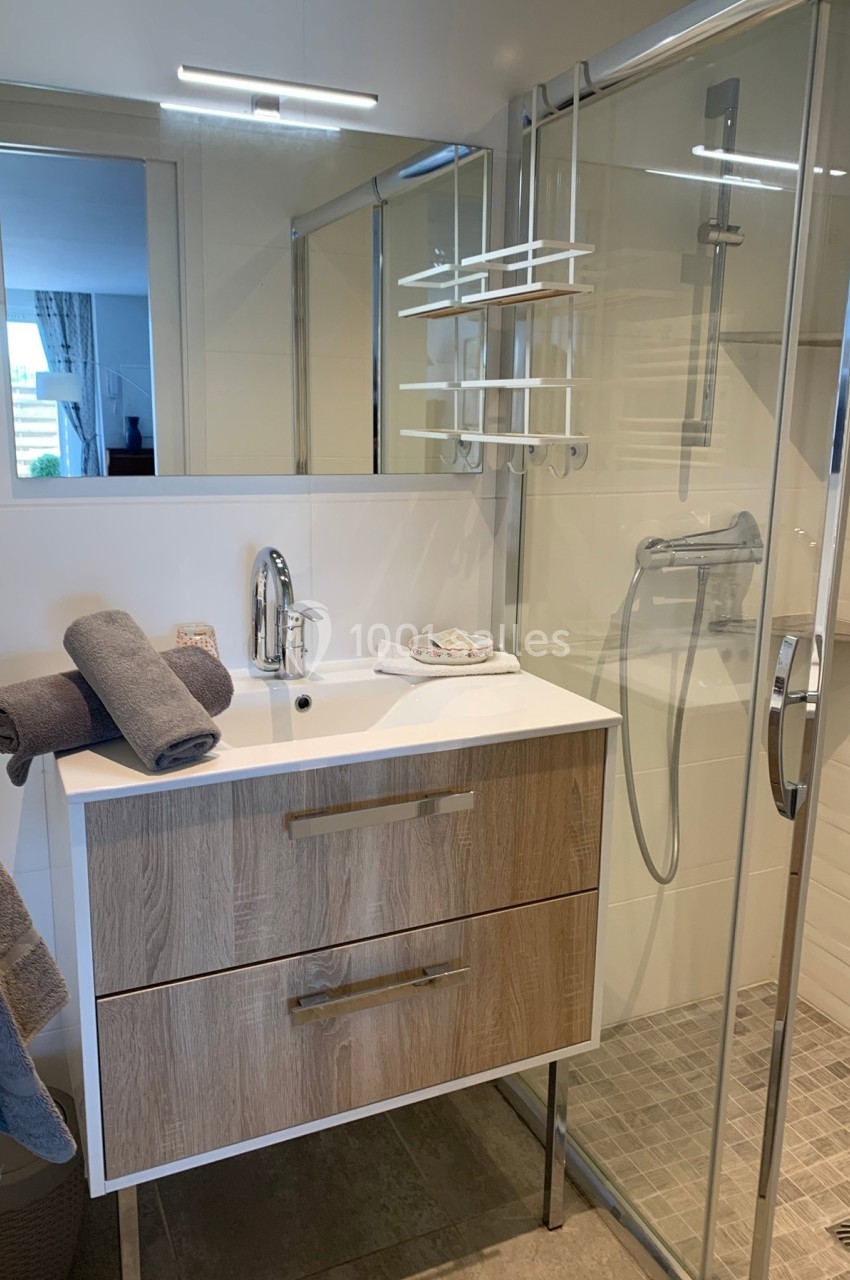Salle de bain moderne avec meuble en bois, lavabo, miroir, douche vitrée et serviettes grises pliées.