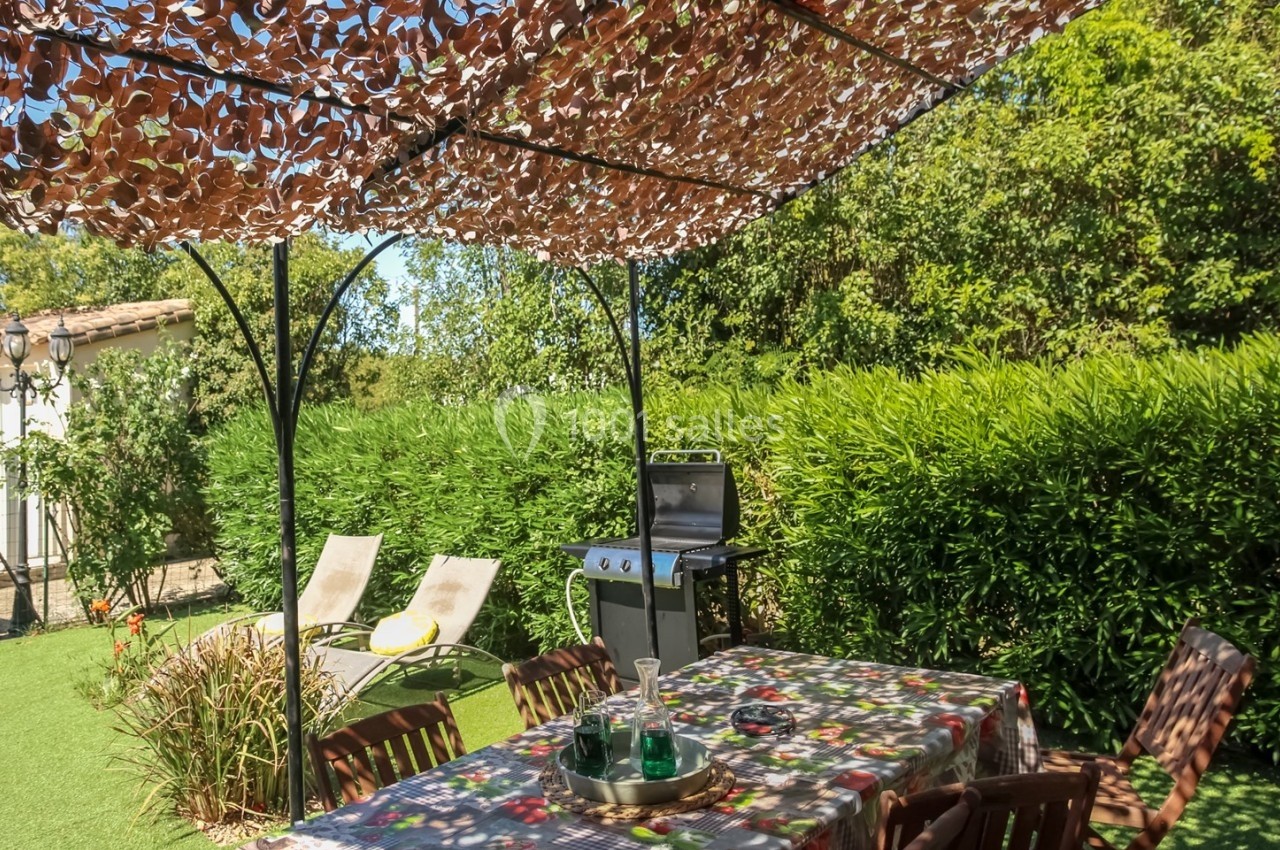 Table de jardin avec nappe colorée sous une tonnelle ombragée, barbecue et transats dans un jardin verdoyant.
