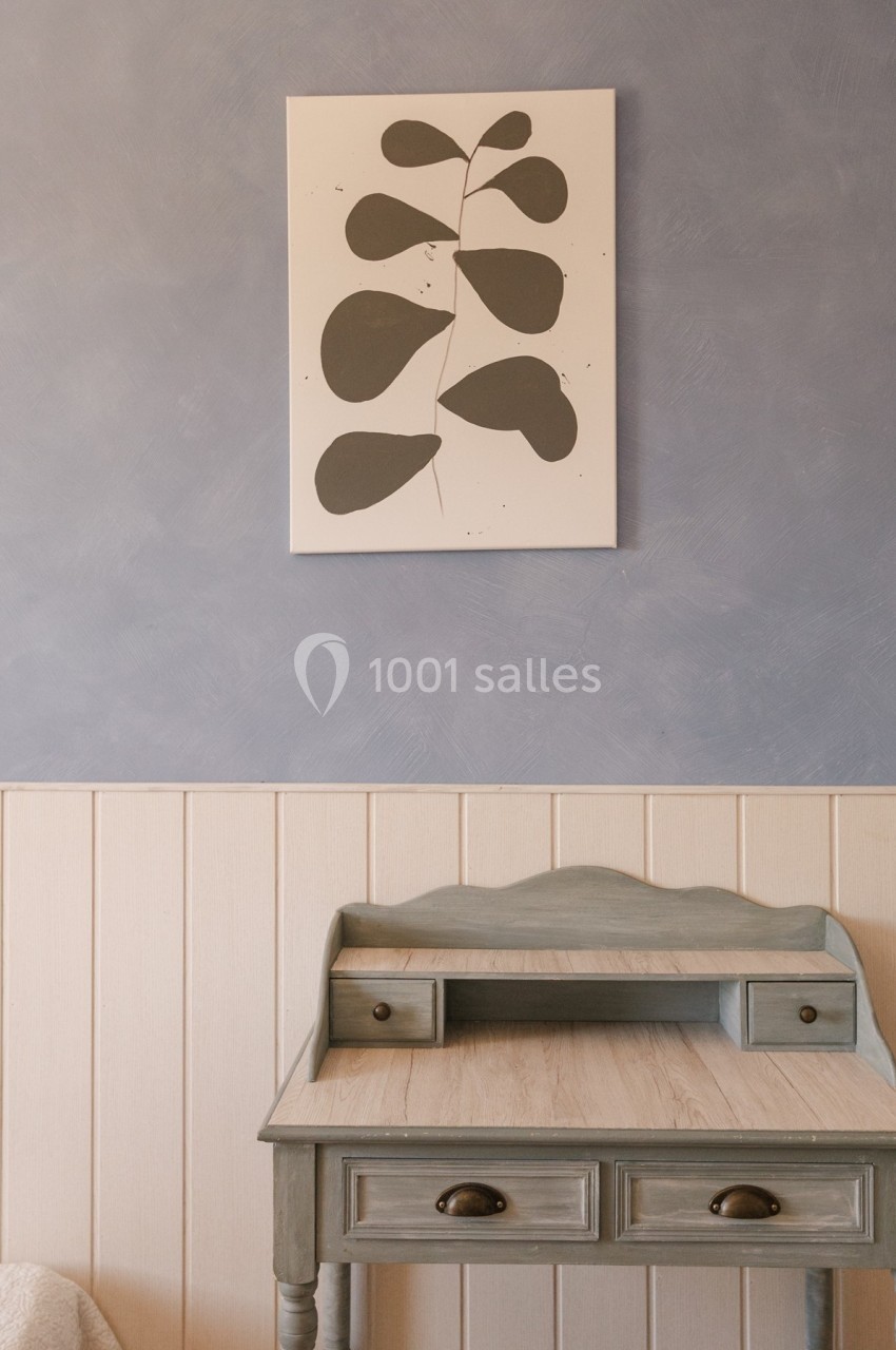 Table en bois avec tiroirs devant un mur bleu et beige, décoré d'un tableau minimaliste représentant des feuilles.