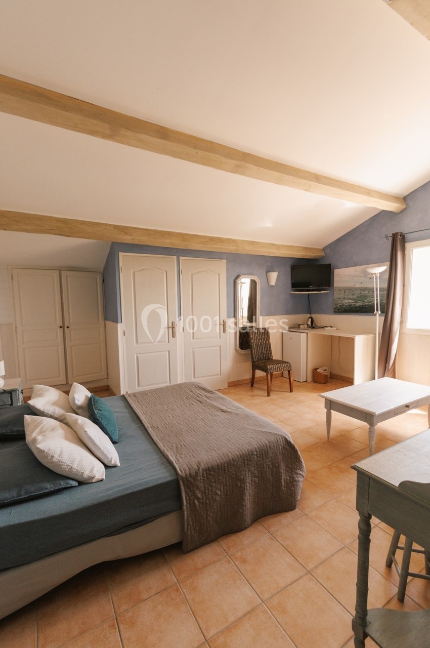 Chambre lumineuse avec lit double, bureau, chaise, armoire, et poutres apparentes sous un plafond incliné.