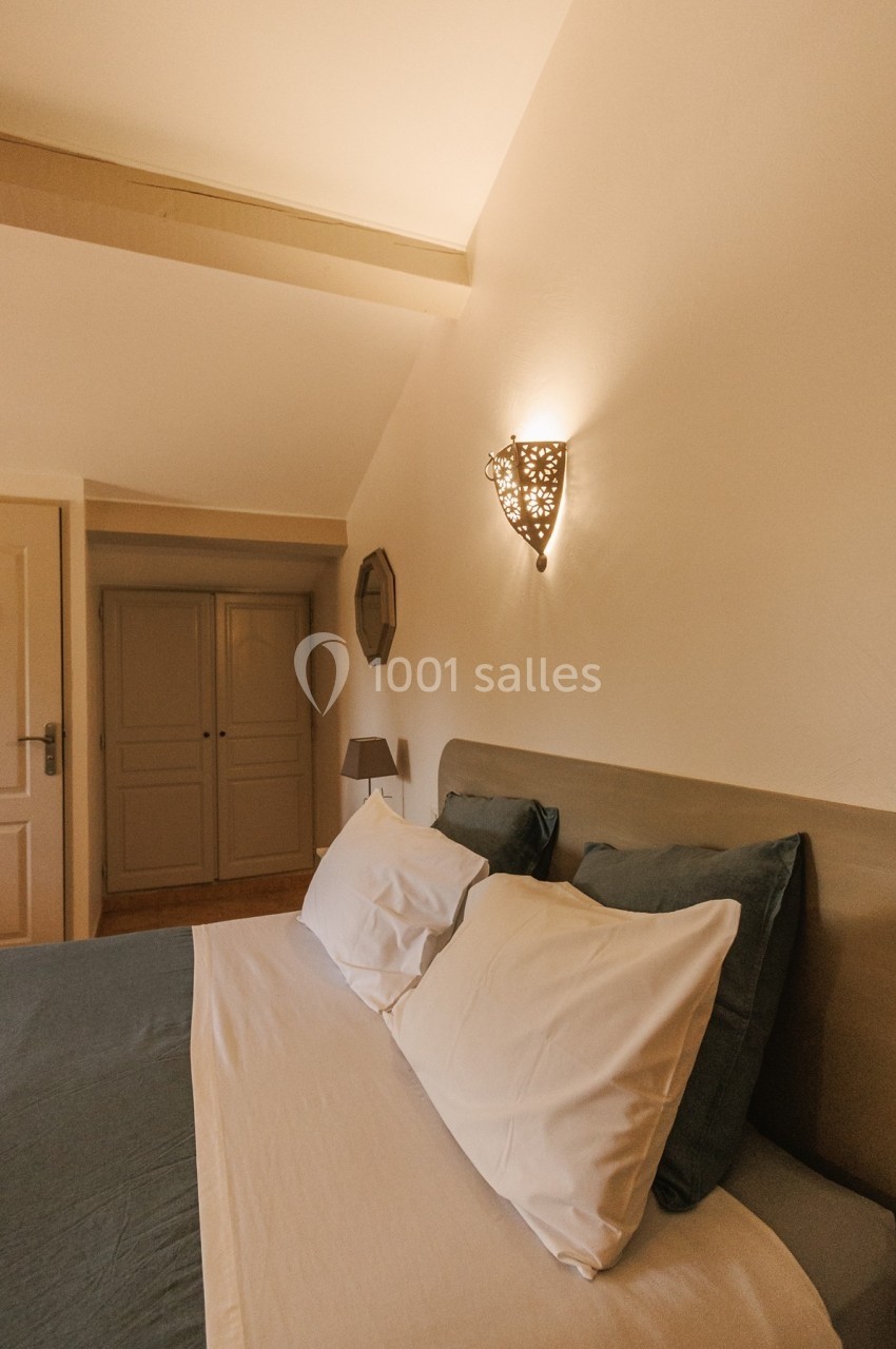 Lit double avec draps blancs et coussins verts dans une chambre lumineuse, éclairée par une applique murale décorative.