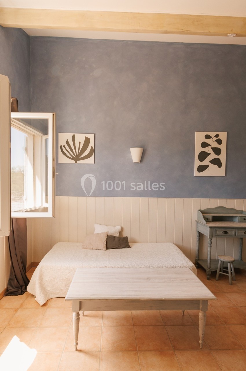 Chambre lumineuse avec un lit simple, table basse, bureau et murs bleu pastel décorés de cadres minimalistes.