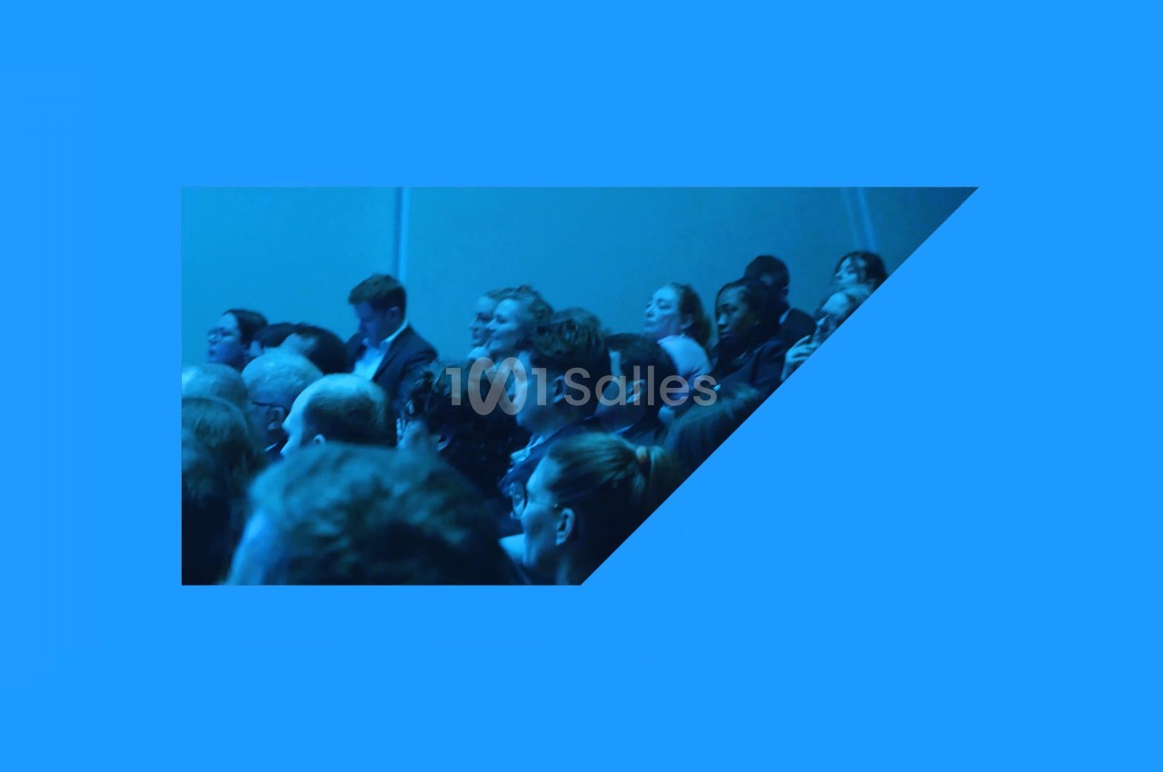 Public assis en rangées, vu de côté, dans une salle baignée d'une lumière bleue.