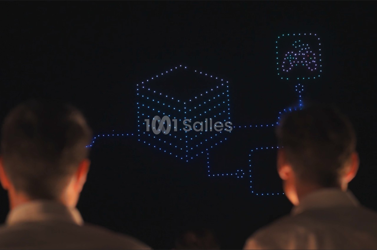 Deux spectateurs de dos regardent un spectacle de drones formant un cube lumineux bleu relié à un pictogramme, de nuit.