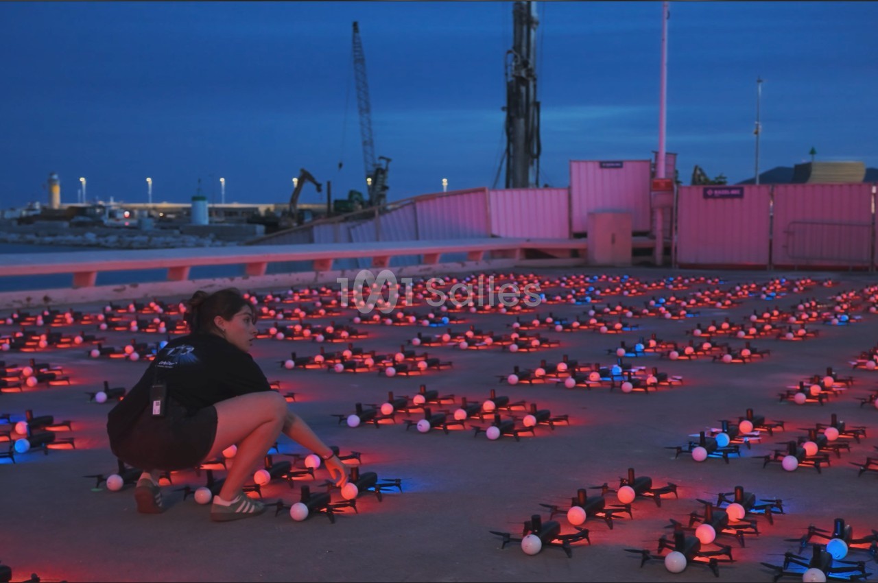Au crépuscule, une femme accroupie regarde des rangées de drones au sol éclairés de lumières rouges et blanches.