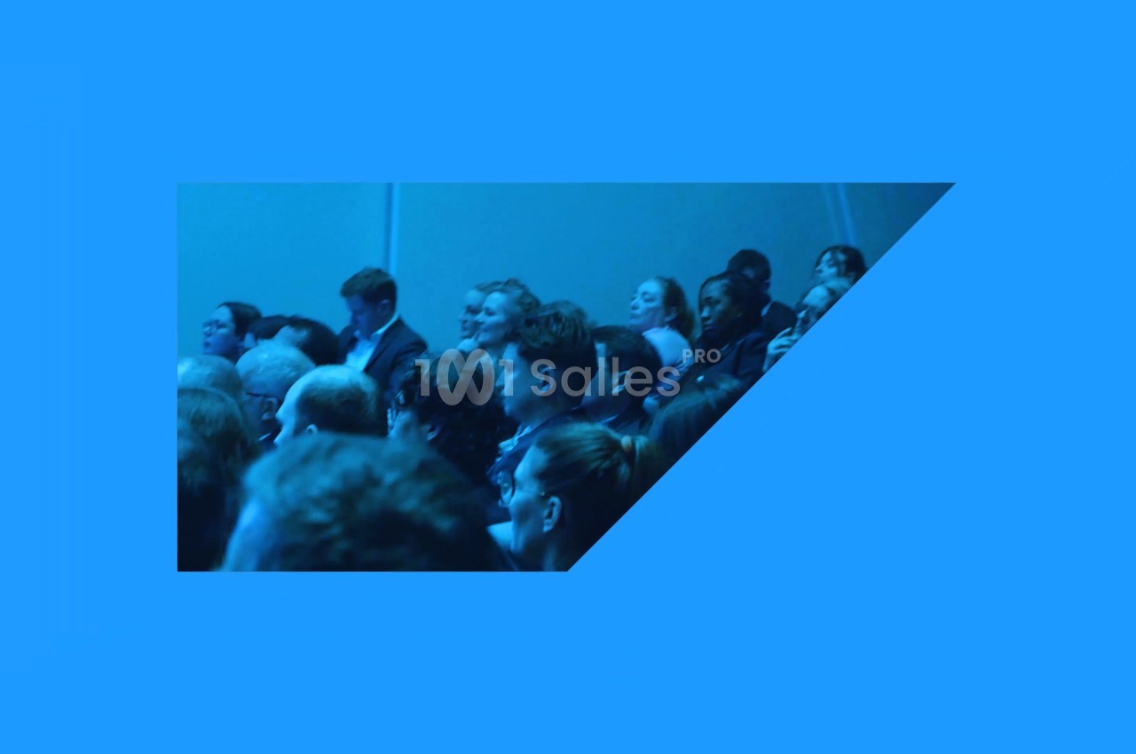 Public assis en rangées, vu de côté, dans une salle baignée d'une lumière bleue.