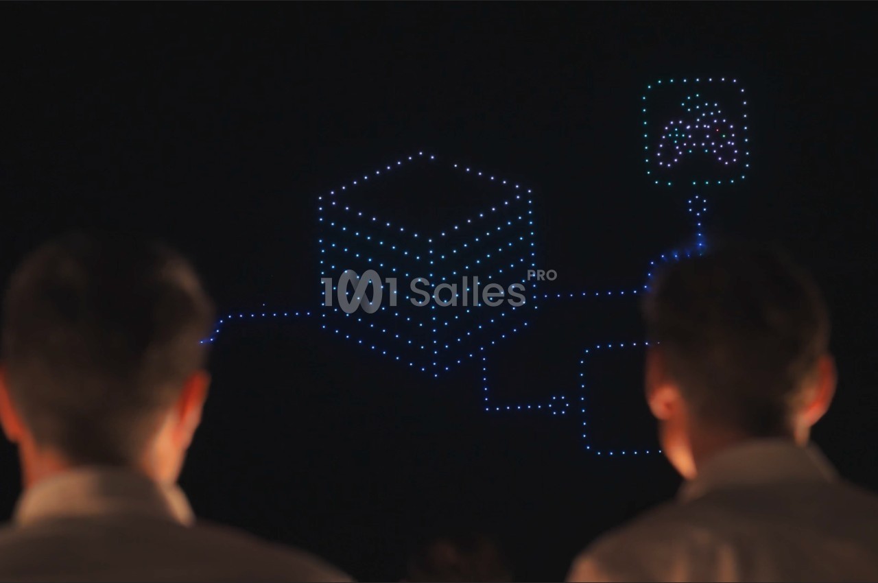 Deux spectateurs de dos regardent un spectacle de drones formant un cube lumineux bleu relié à un pictogramme, de nuit.