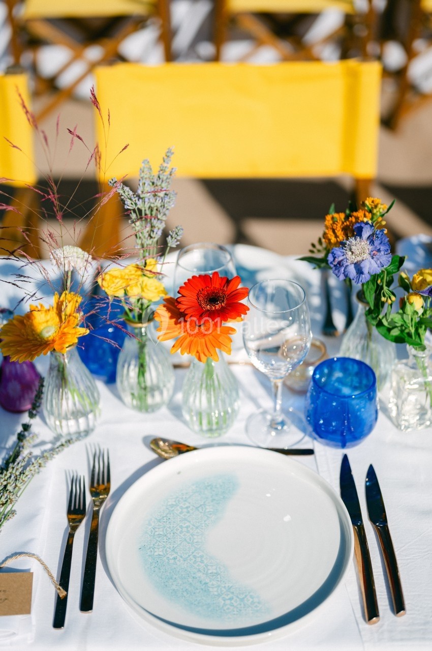 Table dressée avec assiette blanche et bleue, couverts en métal, verres colorés et petits bouquets de fleurs.