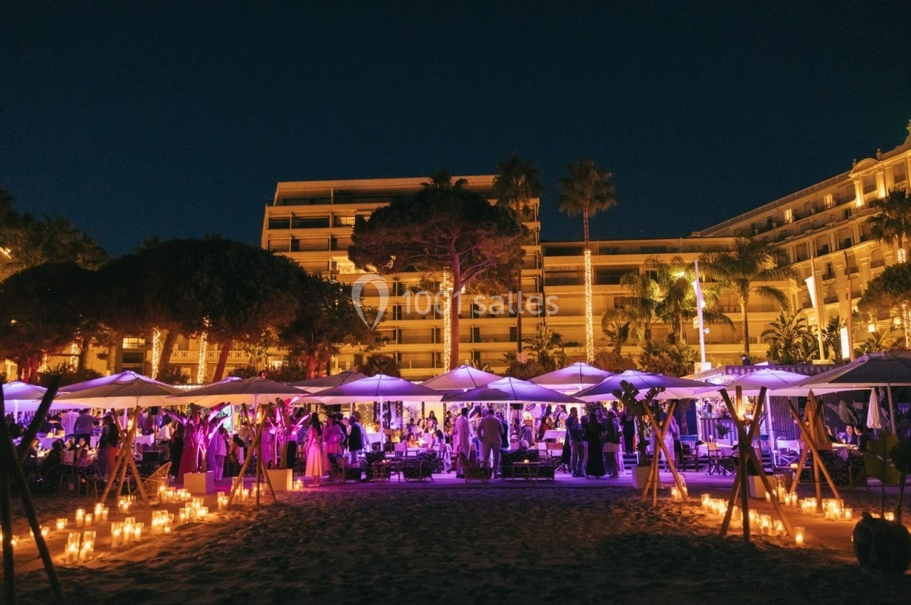 Soirée en plein air sur une plage éclairée par des bougies, avec parasols et un bâtiment illuminé en arrière-plan.