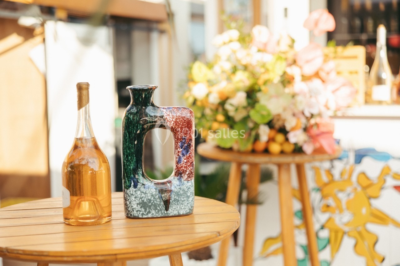 Bouteille de vin et vase coloré posés sur une table en bois, avec un bouquet de fleurs en arrière-plan.