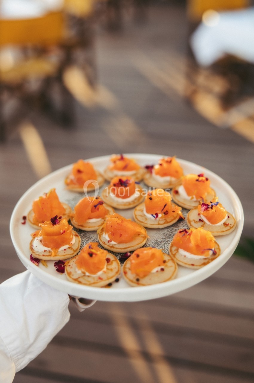 Plateau de blinis garnis de crème, saumon fumé et fleurs comestibles, présenté sur une terrasse ensoleillée.
