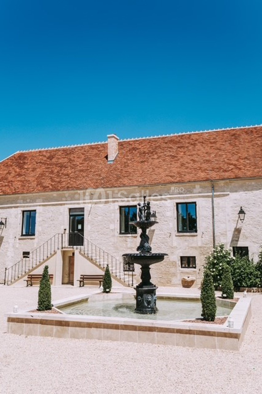 Cour d'une bâtisse en pierre avec toit en tuiles, une fontaine centrale entourée de végétation et ciel bleu.