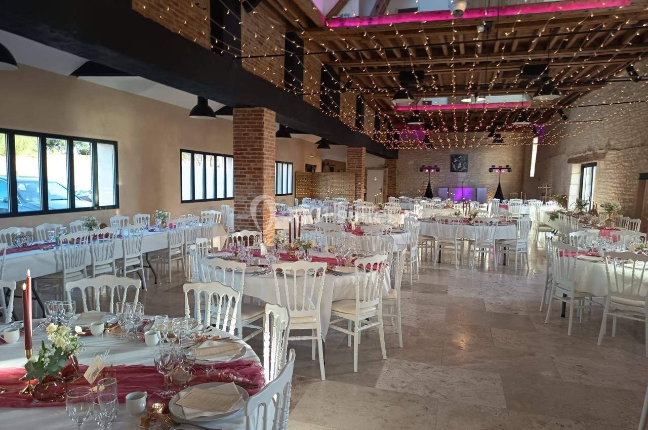Salle de réception décorée pour un événement, avec tables dressées, chaises blanches et guirlandes lumineuses suspendues.