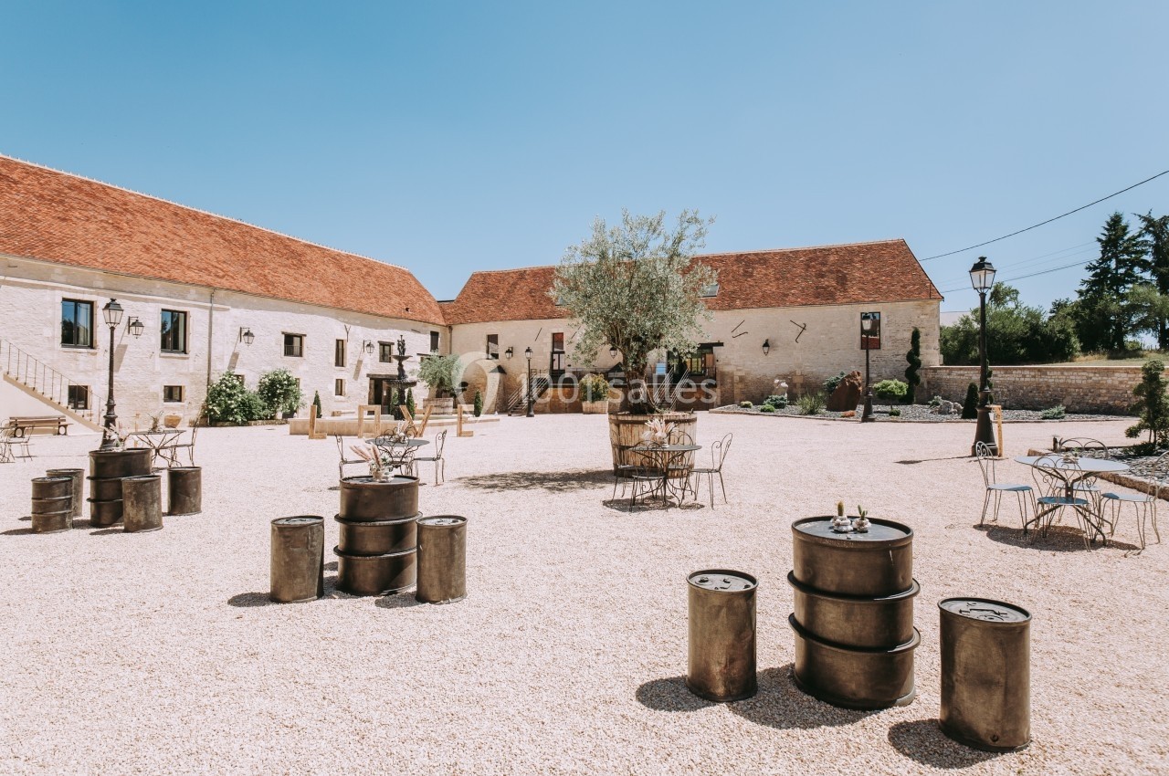 Cour extérieure en gravier avec tables, chaises, tonneaux en métal et bâtiments en pierre sous un ciel dégagé.