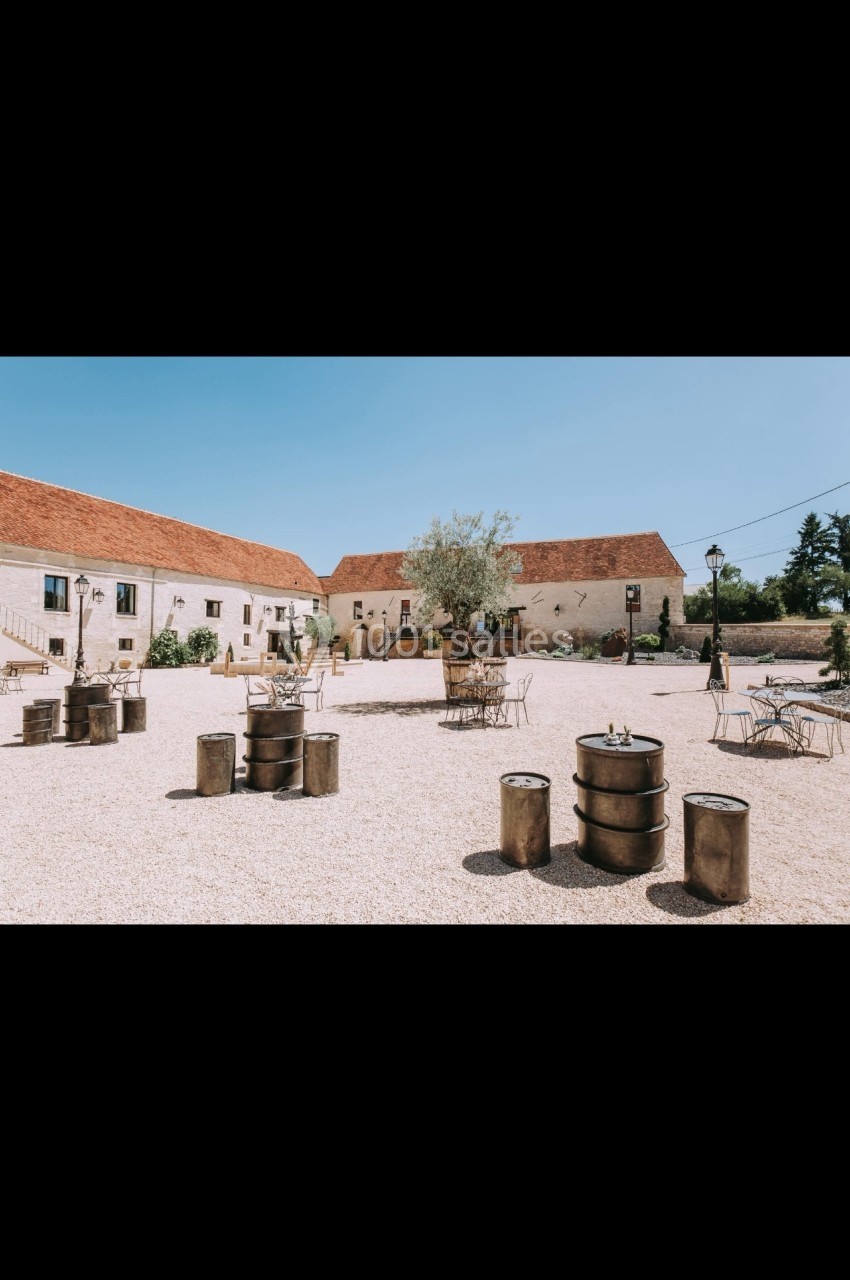 Cour extérieure en gravier avec tables, chaises, tonneaux en bois et bâtiments en pierre sous un ciel dégagé.