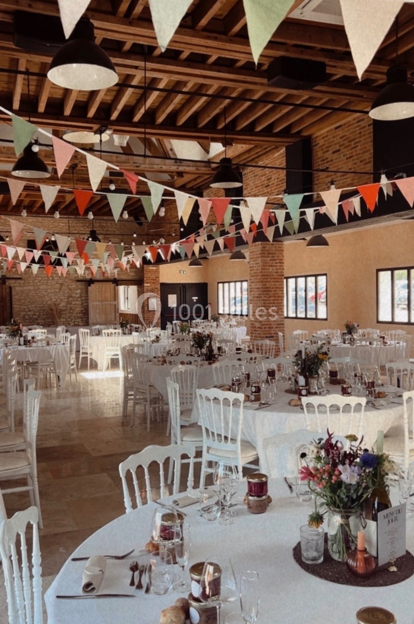 Salle de réception décorée avec des guirlandes colorées, tables rondes dressées et chaises blanches.