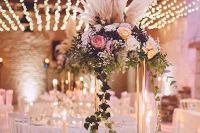 Table en bois avec un chapeau, un flacon de parfum, des sandales, un bouquet de fleurs et une bague devant un mur de briques.