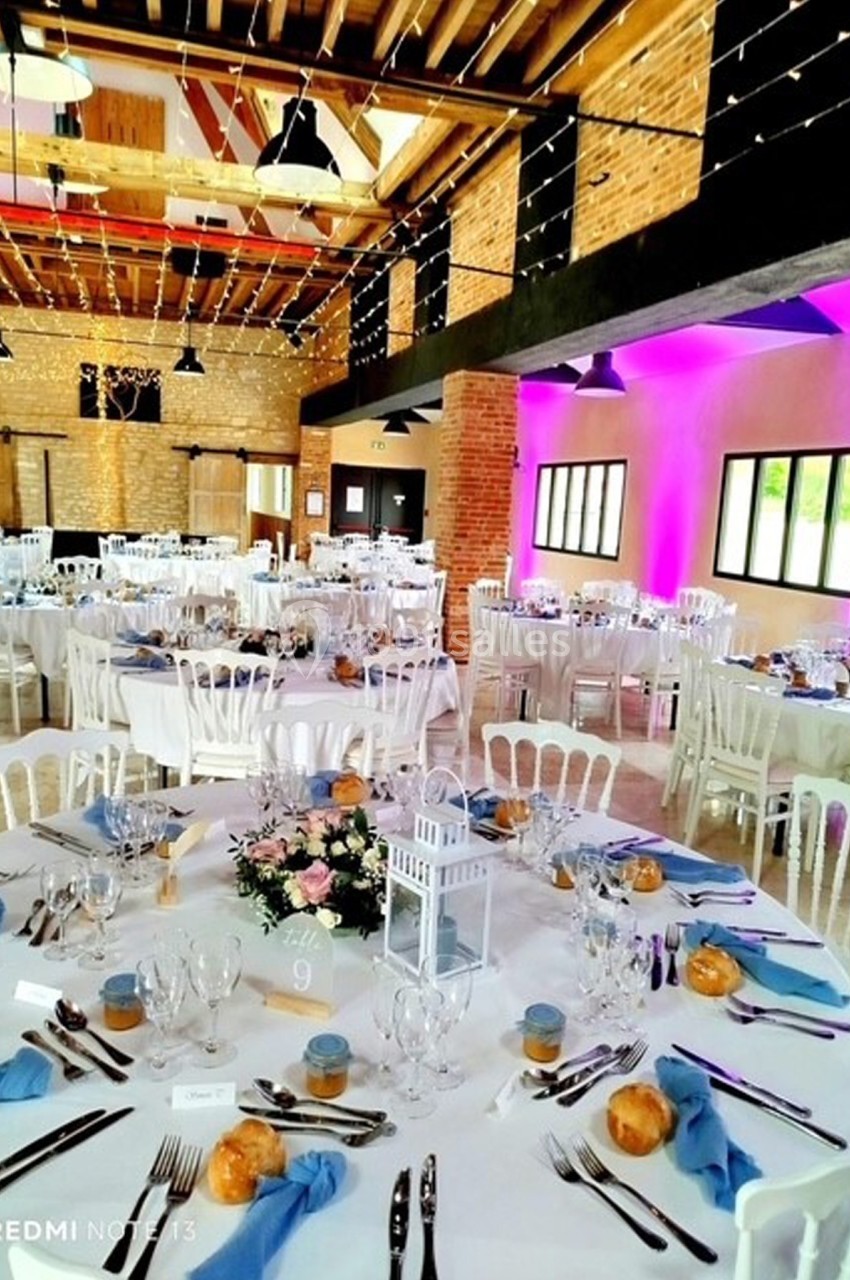 Salle de réception décorée avec des tables rondes dressées, nappes blanches, serviettes bleues et centre de table floral.
