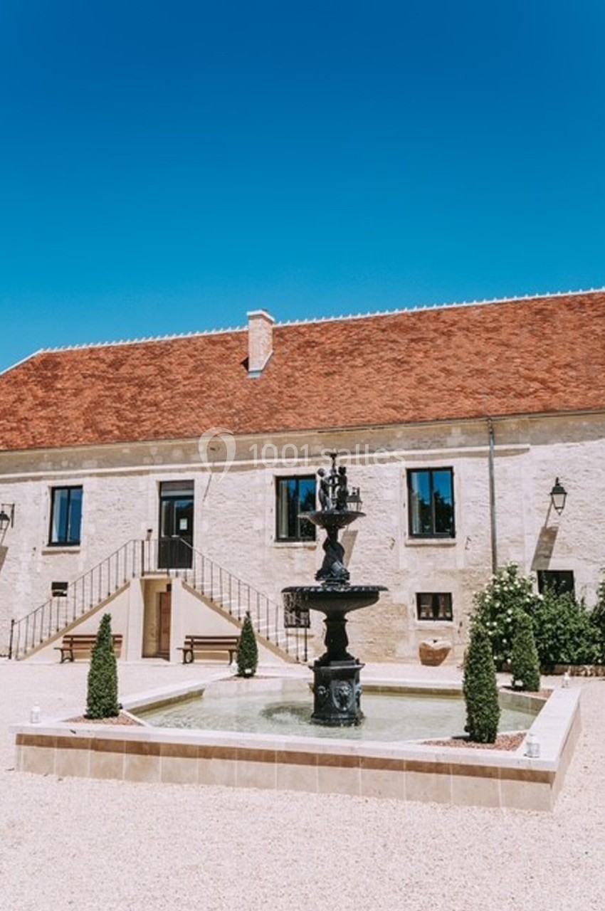 Cour pavée avec une fontaine centrale, entourée d'un bâtiment en pierre à toit rouge sous un ciel bleu clair.