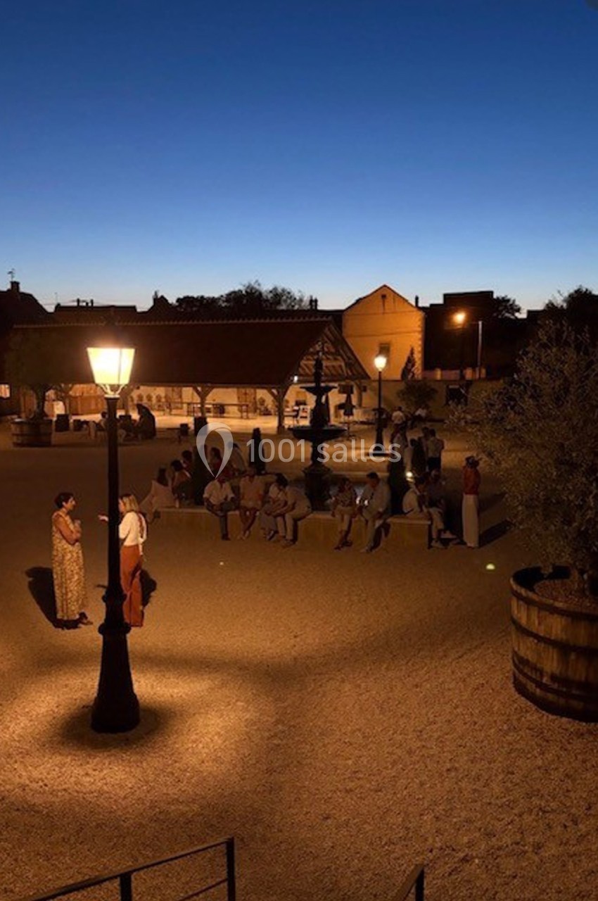 Place éclairée par des lampadaires au crépuscule, avec des personnes rassemblées près d'une structure en bois.