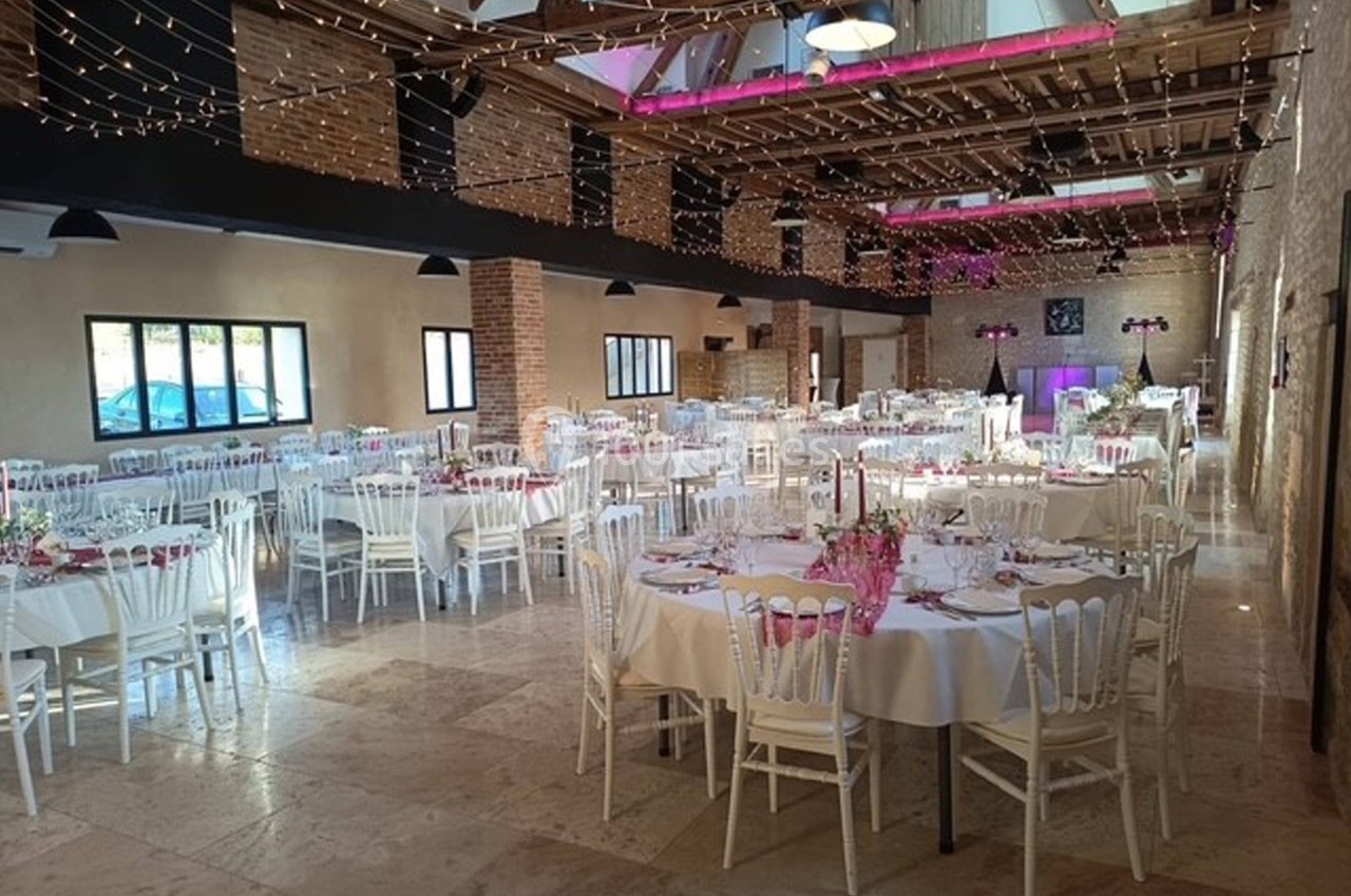 Salle de réception décorée avec des tables rondes, nappes blanches, chaises élégantes et guirlandes lumineuses suspendues.