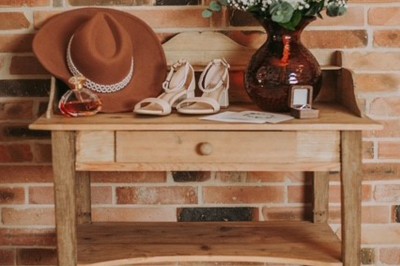 Table en bois avec un chapeau, un flacon de parfum, des sandales, un bouquet de fleurs et une bague devant un mur de briques.