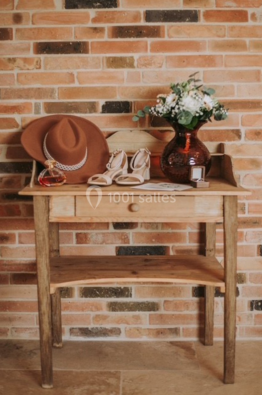 Table en bois avec un chapeau, un flacon de parfum, des sandales, un bouquet de fleurs et une bague devant un mur de briques.