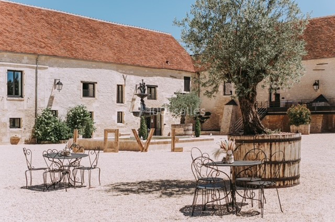 Cour d'une bâtisse en pierre avec tables en fer forgé, un olivier dans un tonneau et des lettres formant ’LOVE’.