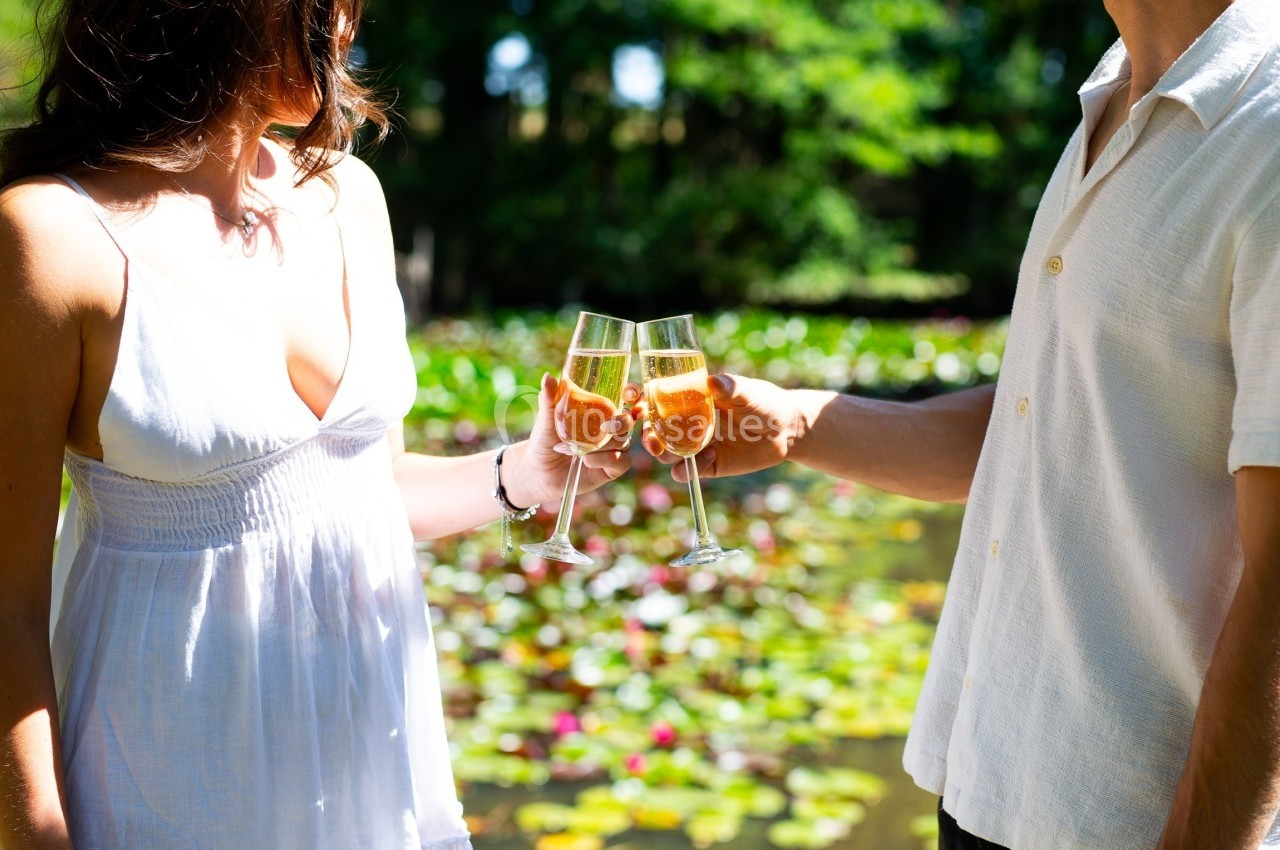 Deux personnes trinquent avec des verres de champagne devant un étang couvert de nénuphars, en plein jour.