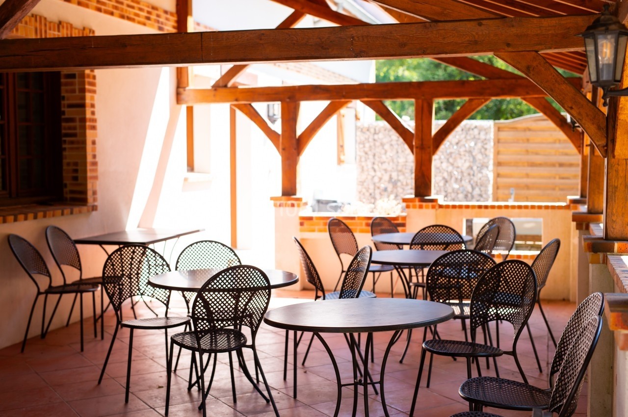 Terrasse couverte avec des tables et chaises en métal noir, entourée de structures en bois et murs en briques.