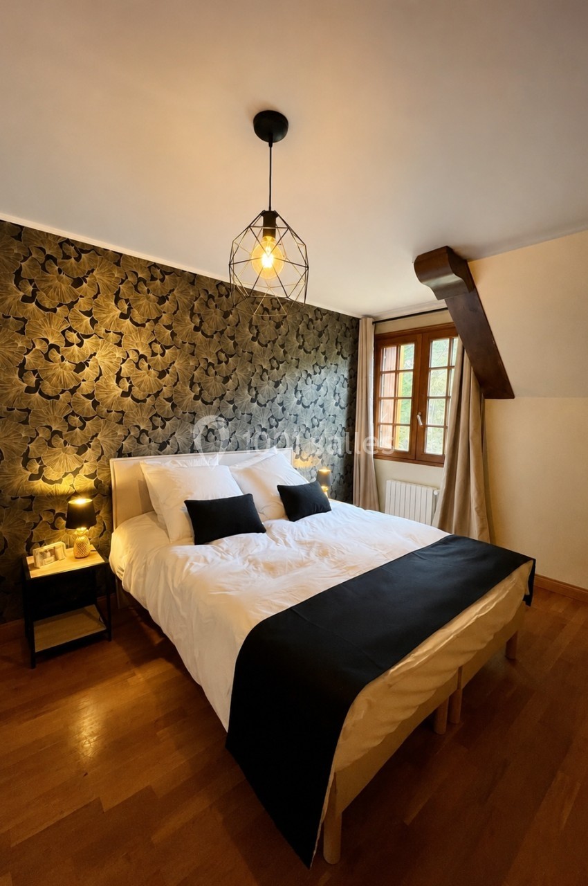 Chambre avec lit double, linge blanc, coussins noirs, mur à motif floral sombre, parquet et fenêtre en bois.